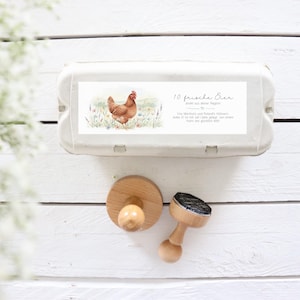 May include: A white egg carton with a label featuring a watercolor illustration of a brown hen in a field of flowers. The label reads "10 fresh eggs" and includes additional text. Two wooden stamps are also visible.