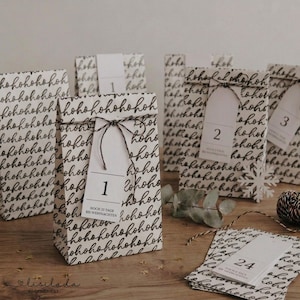 May include: A set of white paper bags featuring a repeating black 'hohoho' pattern. Each bag is tied with black and white twine and includes a white rectangular tag with a number and the German text 'NOCH 23 TAGE BIS WEIHNACHTEN'.