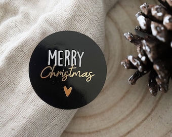 Puede incluir: Pegatina redonda negra con el texto "Merry Christmas" en blanco y un corazón dorado. La pegatina está sobre un fondo de tela beige.