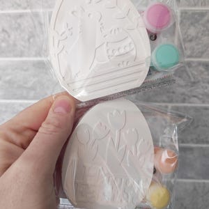 May include: Two Easter egg-shaped craft kits in clear packaging. One egg features a bunny and floral design, the other says "Happy Easter" with tulip designs. Each kit includes paint pots in pink, turquoise, orange, and yellow.