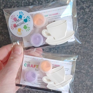 May include: Two craft kits for painting a teacup and saucer. Each kit includes a small white teacup and saucer, a small pot of orange paint, a small pot of purple paint, and a small paintbrush. The kits are packaged in clear plastic bags with a small card that says "little CRAFT hut" and "NAP TAG SHARE".