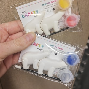 May include: Two clear plastic packages each containing two white polar bear figurines and three small paint pots with yellow, red, blue, and clear paint. The packages have the text "little CRAFTY hugs" and "SNAP TAG SHARE".