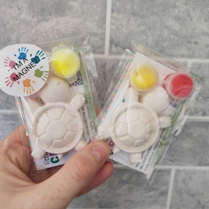 May include: Two white ceramic turtle figurines with a magnet on the back. Each turtle comes with a small bottle of yellow paint, a small bottle of red paint, and a small bottle of white paint. The packaging includes the text "I'm a Magnet".