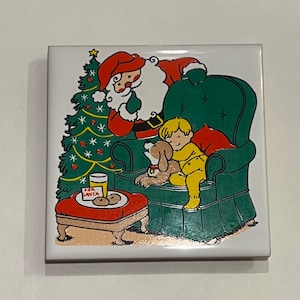 May include: A ceramic tile with a Christmas scene featuring Santa Claus, a child, and a dog. Santa is sitting in a green armchair, looking at the child who is sleeping with a dog in their arms. There is a Christmas tree in the background and a plate of cookies on a small table with the text "For Santa".