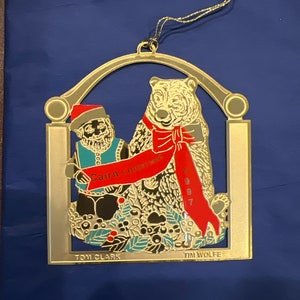 May include: Gold-coloured metal Christmas ornament featuring a bear and a small figure wearing a Santa hat. The ornament has the text "Cairn Christmas 1997" and the names "Tom Clark" and "Tim Wolfe".