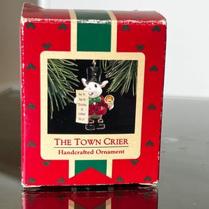 May include: A red box with a green stripe and a framed image of a white mouse in a top hat holding a sign that says "Christmas Joy is Always Near Ye!". The box reads "The Town Crier Handcrafted Ornament".