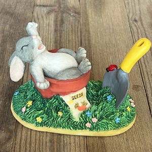 May include: A gray and white rabbit figurine is lying in a red flower pot with a yellow and green 'SEEDS' label. A small red ladybug is on a gray miniature shovel with a yellow handle.