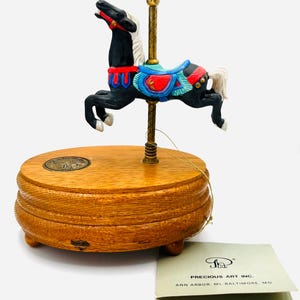May include: A miniature carousel horse figurine in black with a white mane and tail, adorned with a red, blue, and black saddle. The horse is mounted on a brass pole atop a wooden base. A tag from Precious Art Inc. is visible.