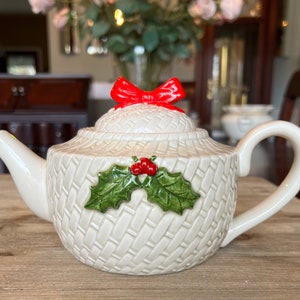 May include: White ceramic teapot with a woven pattern and a red bow on the lid. The teapot has a holly and berry design on the front.