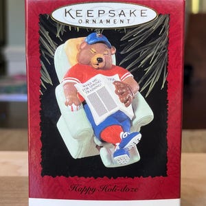 May include: A red box with a white border contains a 1996 Hallmark Keepsake Ornament. The ornament is a brown bear wearing a blue baseball cap, red and blue baseball uniform, and a brown glove. The bear is sitting in a white armchair and reading a newspaper with the headline "Wake Me For Spring Training". The text "Happy Holi-doze" is printed on the box.
