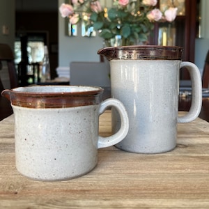 May include: A set of two ceramic jugs with a speckled glaze and brown trim. The larger jug has a handle and a spout. The smaller jug has a handle. Both jugs are on a wooden surface.