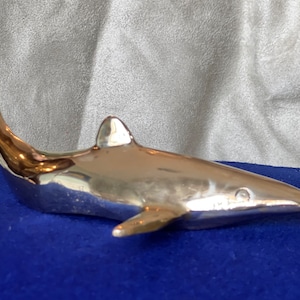 May include: A silver-colored metal sculpture of a shark. The shark is in a swimming position with its tail raised and its head tilted slightly to the side. The sculpture is on a blue surface.
