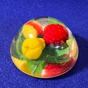 May include: A clear glass paperweight with a red, yellow, and green fruit arrangement inside. The fruit is made of plastic and looks like a cherry, a pear, and a peach.