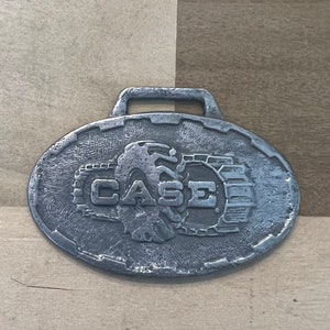 May include: A silver metal keychain with an oval-shaped charm featuring the CASE logo. The logo depicts a stylised tractor with the word CASE in bold letters.