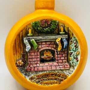 Könnte beinhalten: Eine runde, gelbe Weihnachtskugel mit einer detaillierten Innenszene. Die Szene zeigt einen Ziegelkamin mit Strümpfen, einen Kranz und einen Weihnachtsbaum. Der Schmuck hat eine Metallschlaufe zum Aufhängen.