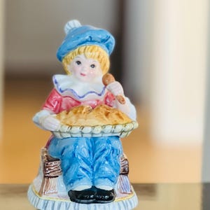 May include: A ceramic figurine of a seated figure in blue pants, a red top, and a blue hat, holding a pie. The figurine has blonde hair and a white ruffled collar. The pie is golden brown and sits in a decorative basket.
