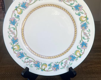 May include: White ceramic plate with a floral design. The plate features a gold border and a decorative pattern of blue and pink flowers with green leaves. The plate is displayed on a black stand.