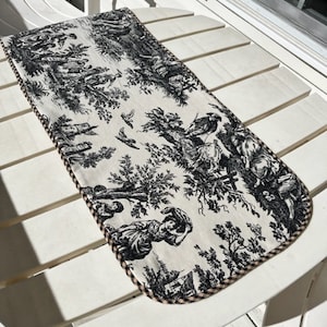 May include: A black and white toile table runner with a black and brown gingham trim. The runner features a repeating pattern of trees, people, and birds.