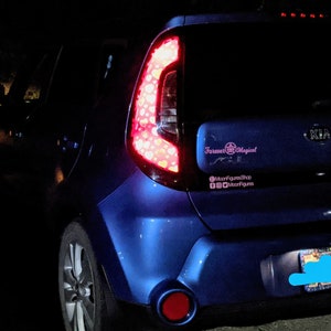 May include: A blue Kia Soul with a pink sticker that says "Forever Magical" and a pink sticker with the text "@MoonfigureShop #Moonfigures". The car has a blue license plate.