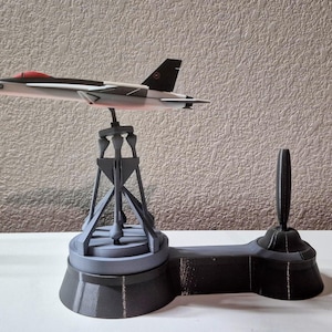 May include: A model airplane, painted in white, black, and red, is mounted on a gray stand. The stand has a circular base and a support structure. A black control stick is attached to the base.