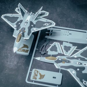 May include: A model kit of an F-22 Raptor fighter jet, featuring a gray and black camouflage pattern. The kit includes the assembled jet on a stand, and unassembled parts on a sprue. The stand has the text "F22 RAPTOR".