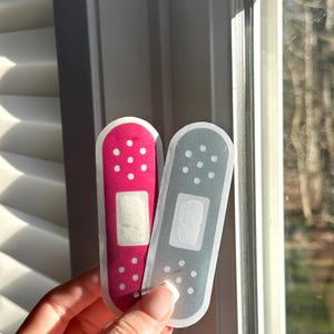 May include: Two adhesive bandages, one pink and one gray, with white accents. The bandages are on a window.