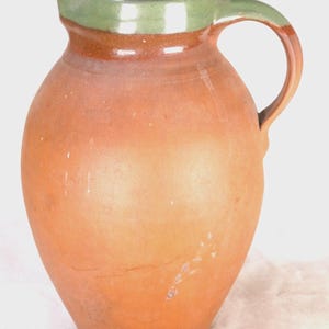 May include: A rustic, earthenware pitcher with a rounded body and a handle. The pitcher is primarily terracotta in color, with a green band around the rim and a dark brown interior. The handle is also green and brown.