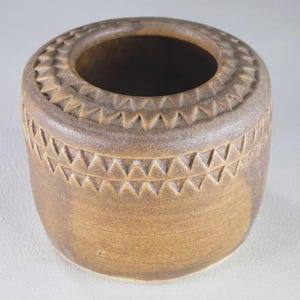 May include: A cylindrical brown ceramic pot with a wide circular opening. The exterior surface features two horizontal bands of incised triangular patterns encircling the upper portion of the vessel, with a smooth finish on the lower body.