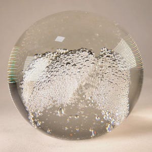 Puede incluir: Un pisapapeles de vidrio transparente de forma esférica. El interior presenta un cúmulo de pequeñas burbujas, creando un efecto texturizado similar a una nube. El pisapapeles es transparente, permitiendo el paso de la luz.