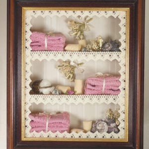 May include: A decorative framed display featuring three shelves. Each shelf holds pink folded towels tied with ribbon, small candles, dried flowers, and other ornaments. The frame is dark brown with a gold inner edge, and the shelves are edged with lace.