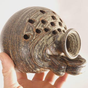 Puede incluir: Escultura de cerámica artesanal con cuerpo bulboso y pico acampanado. La escultura presenta una serie de aberturas circulares y está acabada en tonos marrones y crema, creando un aspecto texturizado y terroso. La pieza tiene una forma única y orgánica.