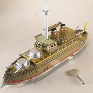 May include: A vintage-style tin toy battleship in gold, featuring two silver-colored gun turrets on the deck, a central mast, two smokestacks with red and black stripes, and a separate silver metal wind-up key for the mechanical movement.