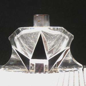 以下が含まれることがあります： 幅の広い角張ったベースと円筒形の金属製キャップを備えた透明なガラス製香水瓶。このボトルは、鋭い幾何学的なファセットと、前面に大胆なコントラストを生み出す目立つ黒い三角形のインレイが特徴です。