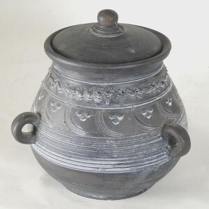May include: A dark grey ceramic jar with a lid and two loop handles. The jar has etched white decorative patterns and horizontal lines. The lid has a small knob. The jar is likely for storage or display.