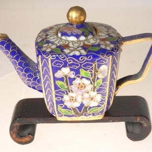 May include: A small, ornate teapot with a blue background and floral designs. The teapot has a gold handle, spout, and lid. It sits on a dark wooden stand. The teapot's design includes white and purple flowers with green leaves.
