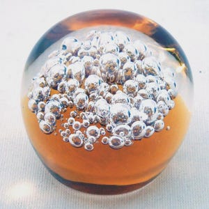 Puede incluir: Un pisapapeles esférico de vidrio transparente con un grupo de burbujas plateadas en su interior. La mitad inferior del pisapapeles tiene un tono ámbar, creando un contraste llamativo. Las burbujas varían en tamaño, agregando interés visual a este artículo decorativo.