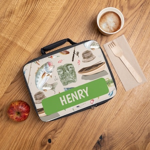 May include: A rectangular lunch bag with a black handle and zipper, featuring a fishing-themed pattern with fish, fishing rods, and a boat. The name "HENRY" is displayed on a green rectangle. An apple, coffee cup, and fork are on a wooden surface.