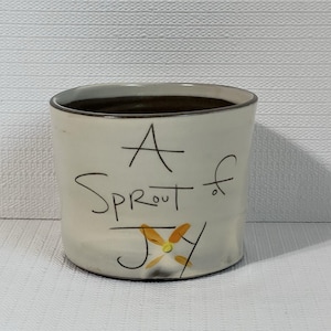 May include: A cylindrical ceramic planter with a cream-colored exterior and a dark brown interior. The planter has the words "A Sprout of Joy" written on the front in black script, with a yellow and orange flower illustration.