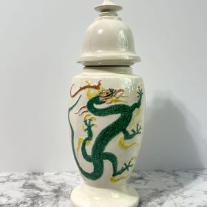 May include: A cream-colored ceramic jar with a lid, featuring a green dragon design with yellow and red accents. The jar has a bulbous body and a pedestal base, suitable for home decor.