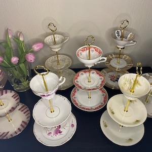 Könnte beinhalten: Ein Set aus drei Keramikbechern mit einer gesprenkelten, cremefarbenen Glasur. Jeder Becher hat ein anderes handgemaltes Design: ein blaues und grünes Blatt, eine gelbe Blume und eine rote Beere. Die Becher haben eine einfache, abgerundete Form.