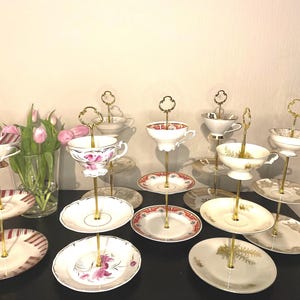 Könnte beinhalten: Ein zweistöckiger Kuchenständer mit goldenen Metallständern und weißen Porzellanplatten. Der Ständer ist mit Vintage-Teetassen und Untertassen mit floralen und goldenen Akzenten verziert.