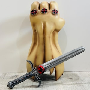 May include: A tan-colored plastic hand with four red and black rings on the fingers. A silver sword with a red and black lion emblem is lying next to the hand.