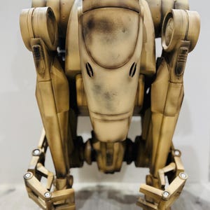 Può includere: Una figura di robot dettagliata, color beige con un aspetto invecchiato. Il robot ha una testa arrotondata con due aperture a forma di occhi e arti articolati. La figura è un oggetto da collezione, forse un modello o un accessorio.