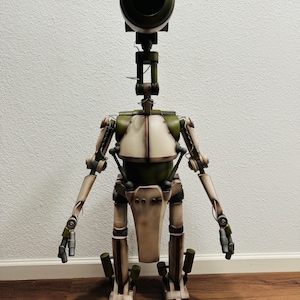 May include: A detailed robot figure with a large, dark green, spherical head and a slender neck. The body is primarily off-white with olive green accents and mechanical details. The figure stands upright with arms at its sides, showcasing a vintage, industrial aesthetic.