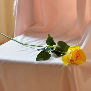 Pode incluir: Uma &uacute;nica rosa amarela com folhas verdes repousa sobre uma toalha de mesa branca.