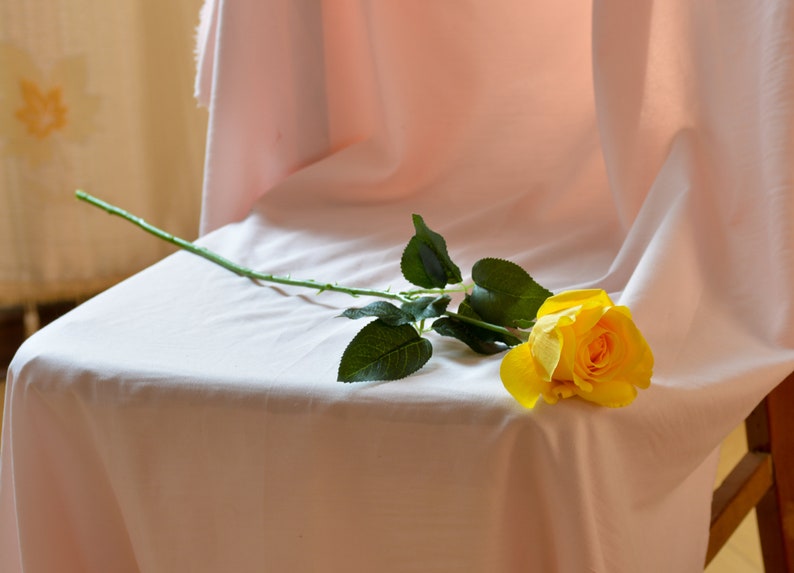 Pode incluir: Uma &uacute;nica rosa amarela com folhas verdes repousa sobre uma toalha de mesa branca.