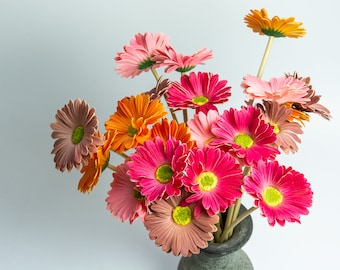 Pode incluir: Um buquê de gérberas coloridas em um vaso cinza. As flores são em tons de rosa, laranja e marrom, com centros amarelos. O arranjo está em um fundo cinza claro.