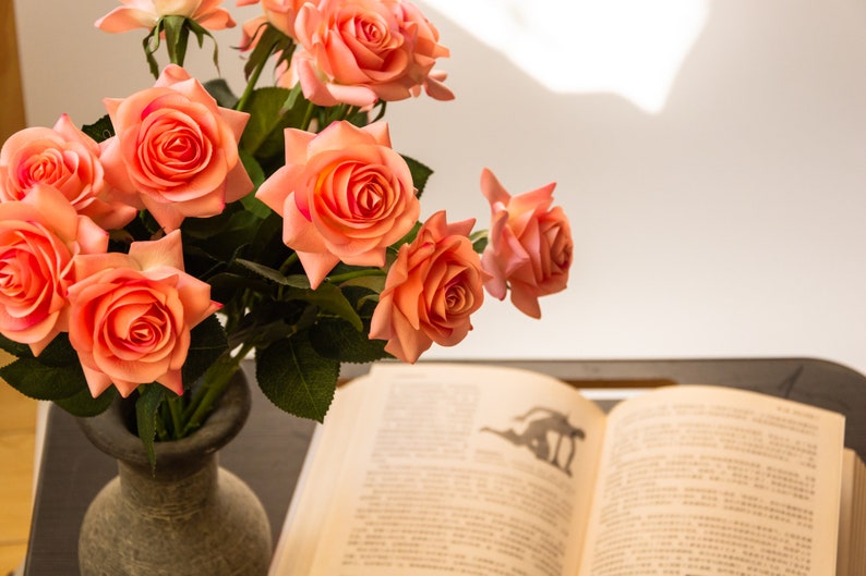 May include: A bouquet of peach-colored roses in a gray stone vase, sitting on a table next to an open book.