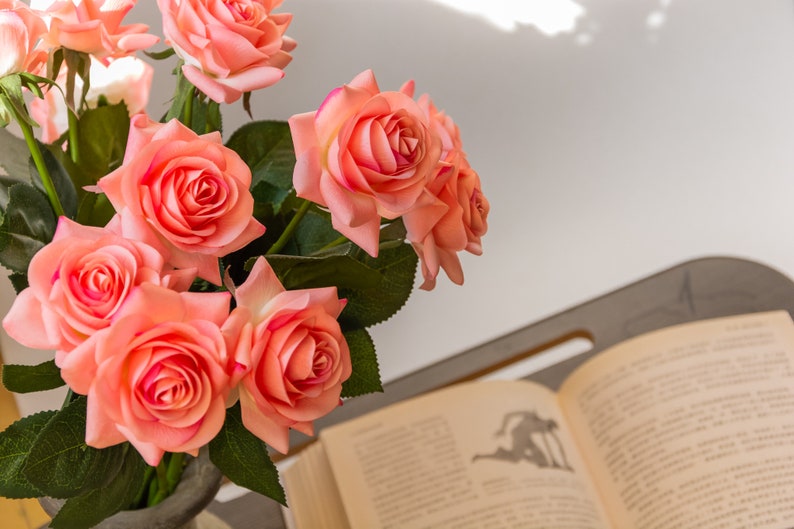 May include: A bouquet of pink roses in a vase, with a book open on a table in the background.