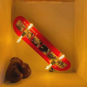 Könnte beinhalten: Rote Skateboard-Lampe mit dem Text "BEASTIE BOYS" und Bildern. Die Lampe hat drei Edison-Glühbirnen. Zwei braune Boxhandschuhe befinden sich in der linken unteren Ecke.